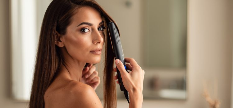 Femme adulte aux cheveux longs, épais et bruns utilisant un lisseur à plaques larges devant un miroir, éclairée par une lumière naturelle douce.