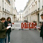 Rue résidentielle française fermée par des barrières municipales, avec trois riverains consultant des documents vierges dans une ambiance de reportage réaliste.